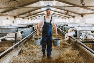 Vista traseira do belo agricultor caucasiano em geral segurando baldes nas mãos com alimentos para animais. Interior estável.