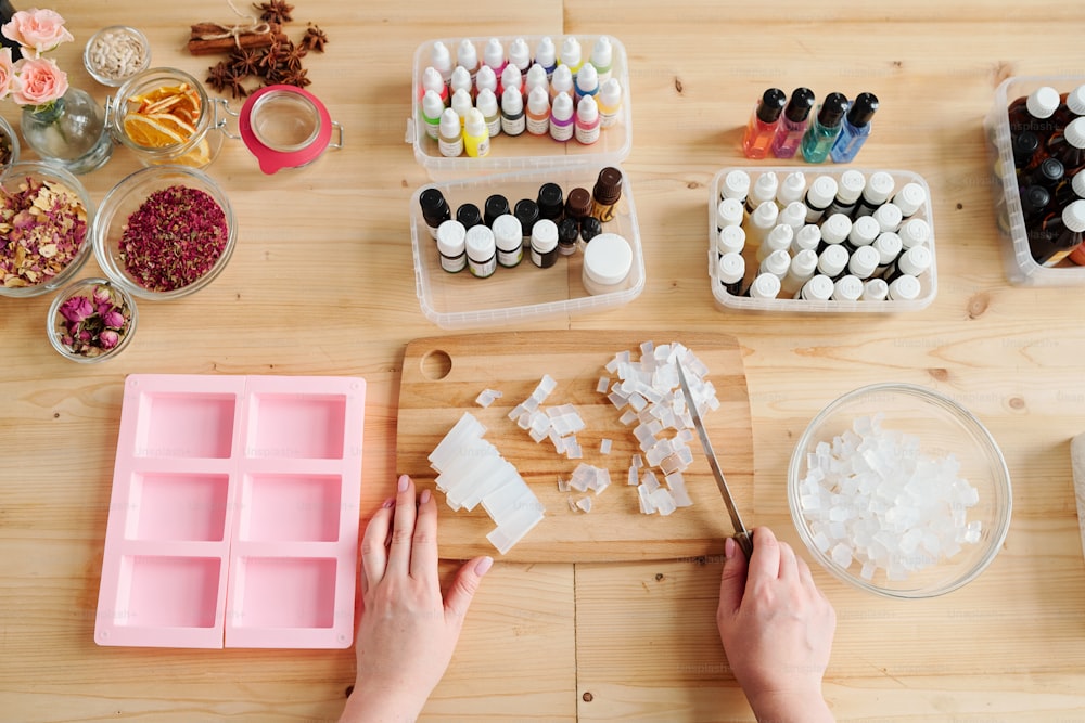 Young woman holding knife over crystals of hard soap mass while cutting it on board among aromatic ingredients