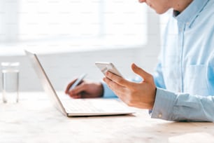 Hand of young man holding smartphone over table while sitting in front of laptop and making working notes