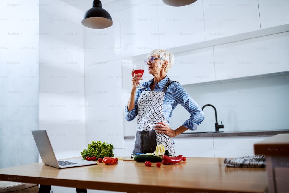 앞치마를 입은 백인 노인 여성은 부엌에 서서 와인을 마시고 건강한 저녁 식사를 준비합니다. 주방 카운터에는 고추, 양상추, 방울 토마토 및 노트북이 있습니다.
