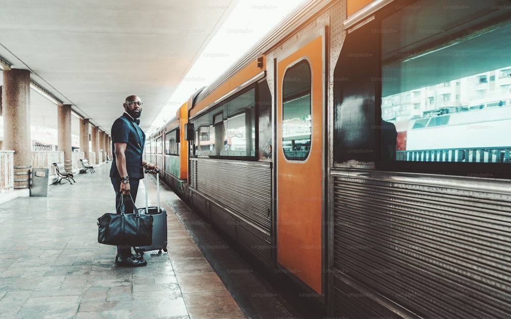 Wide-angle view of an elegant bearded bald mature African man entrepreneur standing with his bags on a railway station platform next to the door of a high-speed train, ready to start his business trip