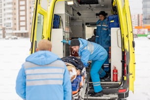Paramedici in abbigliamento da lavoro blu che prendono la barella con un uomo incosciente fissato nell'auto dell'ambulanza prima di andare in ospedale