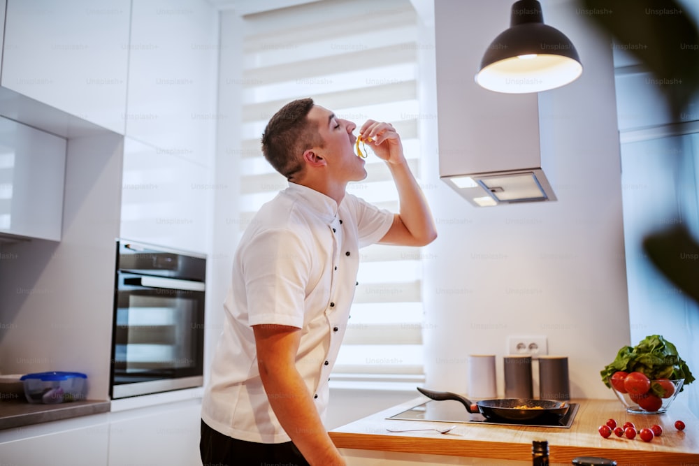 Side view of handsome caucasian chef in white uniform standing in kitchen next to stove and trying fresh pasta. On stove is cooked fresh delicious pasta.