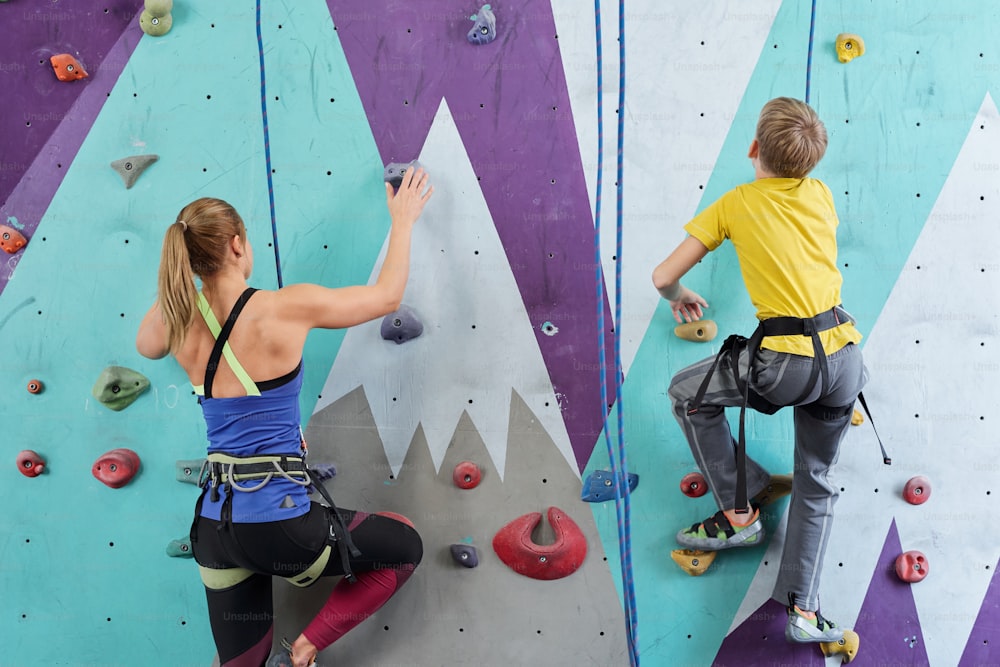Back view of schoolboy and young woman in activewear grabbing by small rocks on climbing wall during sports training