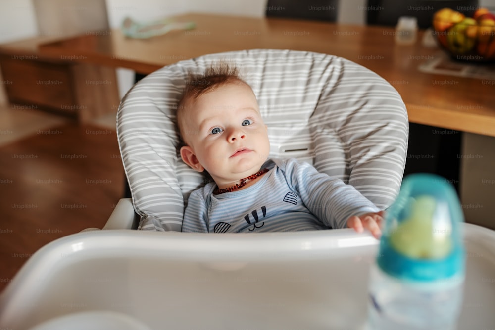Beautiful caucasian curious baby boy sitting in his chair at home and waiting to be feed.
