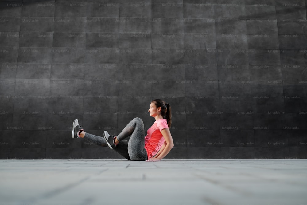 Side view of good-looking brunette in shape and dressed in sportswear sitting on ground outdoors and doing bicycle exercise.