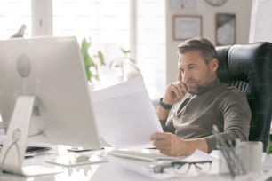 Mature serious architect looking at his sketches while checking them during working day in office