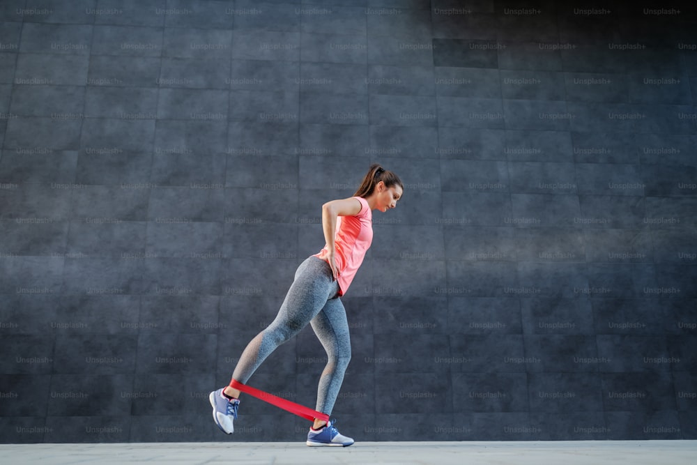Vue latérale d’une charmante brune caucasienne en tenue de sport faisant des exercices pour les jambes avec une bande flexible de pilates.