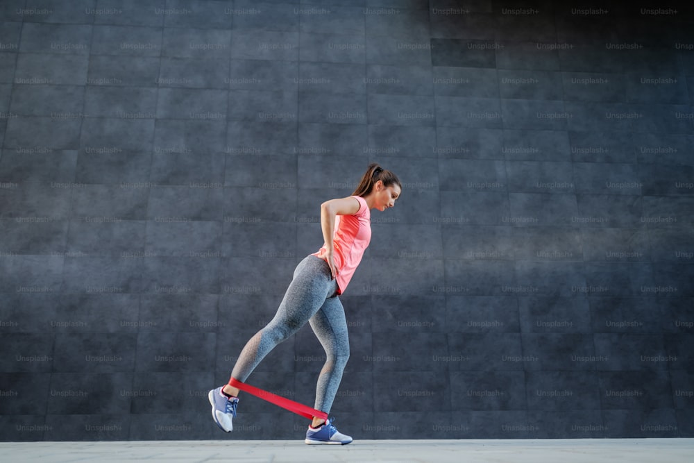 Side view of charming Caucasian brunette in sportswear doing exercises for legs with pilates flexband.