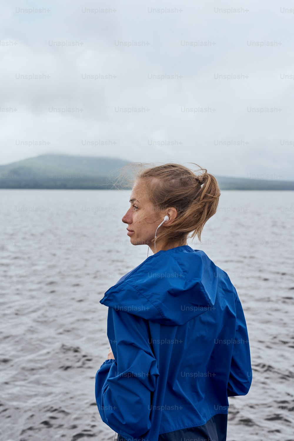 Skinny sportswoman standing outdoor in summer, on gloomy day with scenic view, looking at the lake and mountains, wearing blue rain coat, portrait