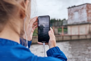 Primer plano de manos femeninas sosteniendo un teléfono inteligente con auriculares enchufados, tomando una foto de un edificio abandonado al aire libre en verano, en un día sombrío con vista panorámica, vistiendo impermeable azul