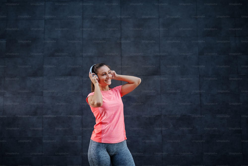 Portrait of attractive smiling Caucasian brunette in sportswear and with ponytail listening music over headphones. In background is dark wall.