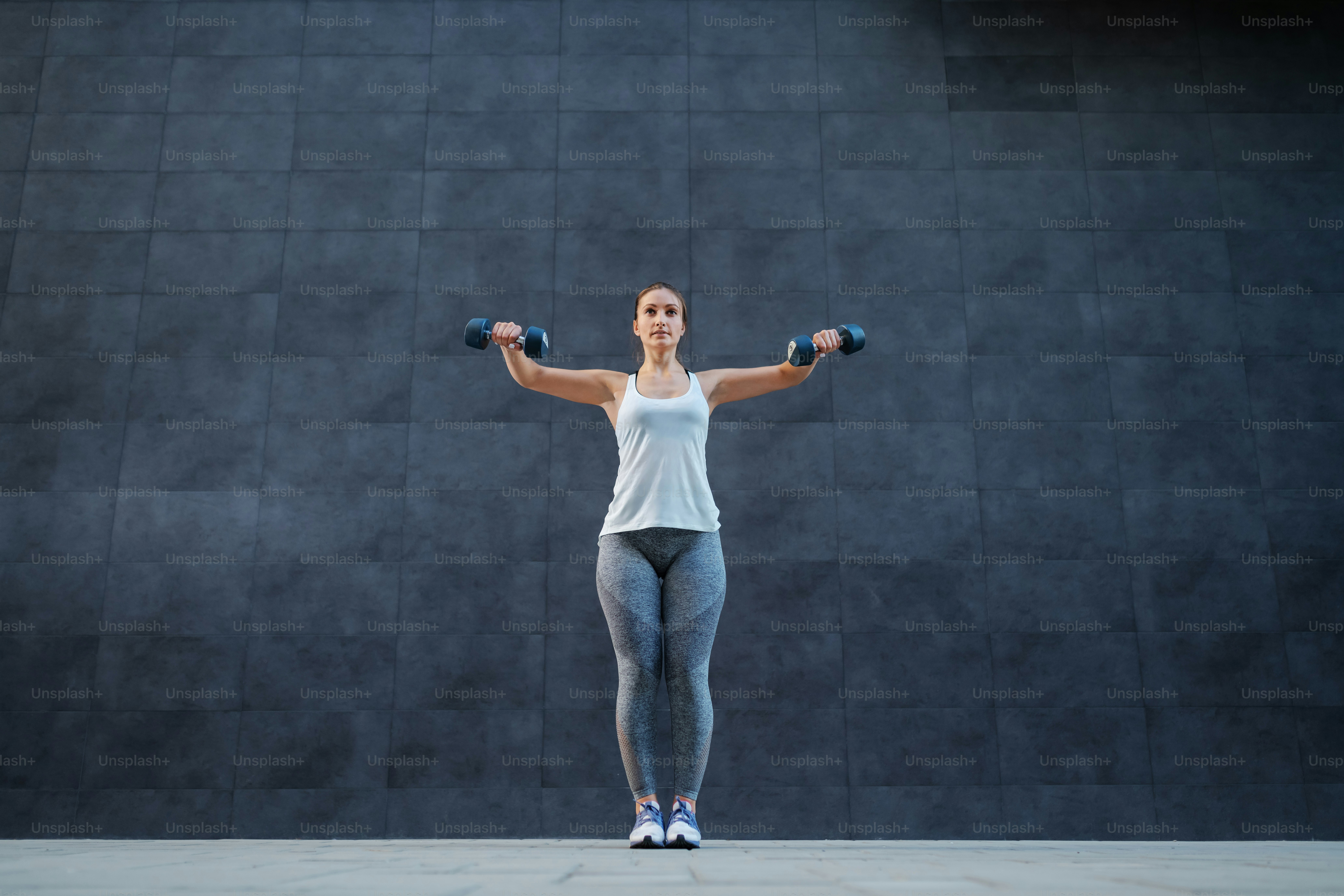 Full length of attractive sportswoman in shape with healthy habits standing outdoors and doing exercises for arms with dumbbells in hands.
