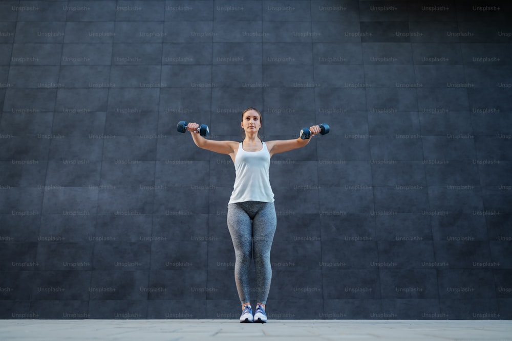 健康的な習慣を持つ形の魅力的なスポーツウーマンのフルレングスは、屋外に立って、手にダンベルを持って腕のエクササイズをしています。