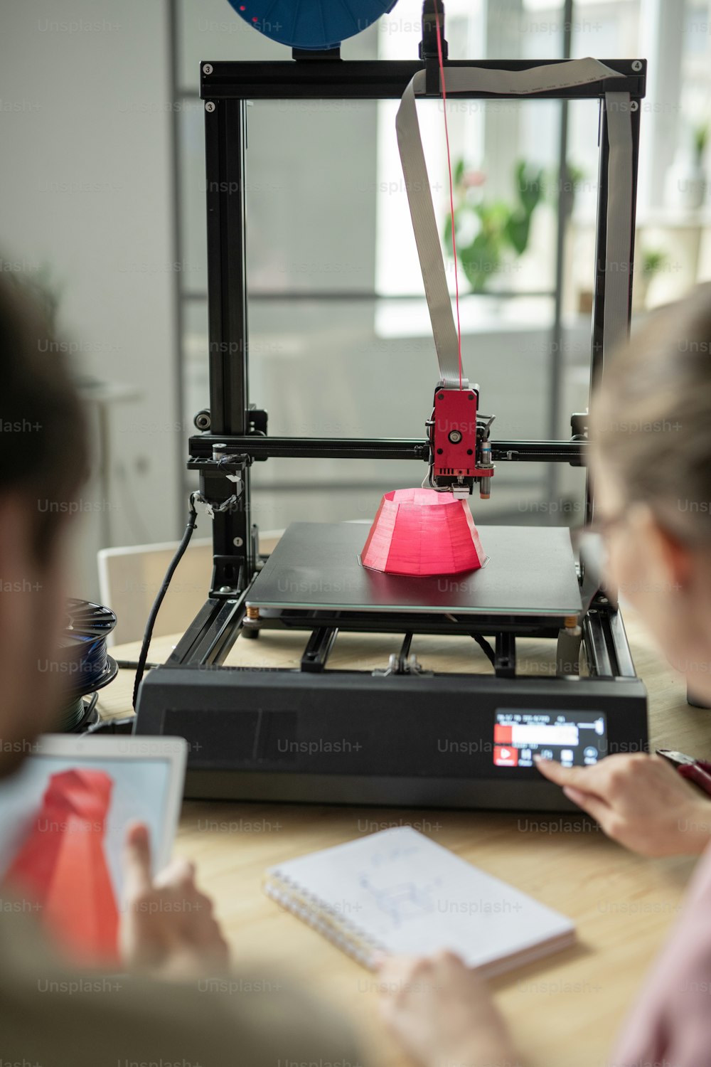 Pink round 3d object is being printed while young woman touching button on control panel of printer
