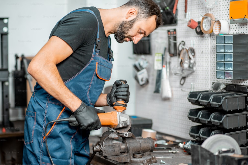 Handyman in overalls cutting metal tube with electric grinder mastering or repairing something in the workshop