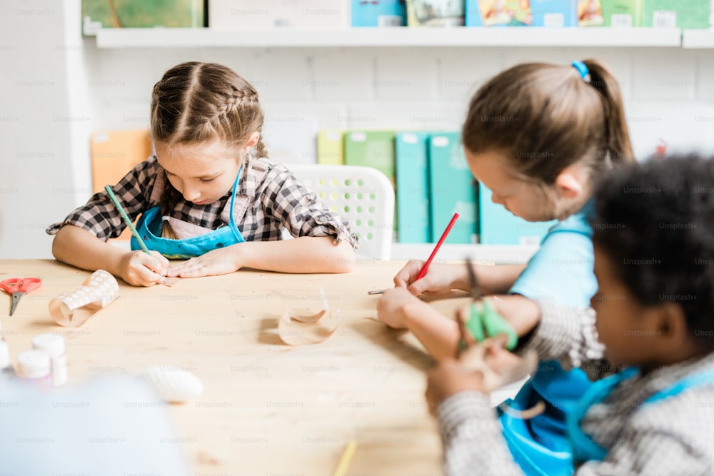 Cute intercultural schoolkids sitting by table and drawing halloween symbols while making decorations for holiday