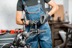 Mechanic in overalls repairing motorcycle, measuring mirror holder for replacement or tuning at the workshop indoors