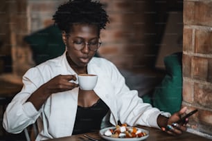 Una joven y encantadora mujer de negocios africana con gafas está bebiendo café caliente mientras está sentada en una cafetería interior y almuerza, con un plato con una deliciosa ensalada frente a ella, un teléfono celular en la mano