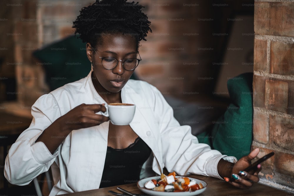 A young charming African business lady in spectacles is drinking hot coffee while sitting in an indoor cafe and having a luncheon, a plate with a delicious salad in front of her, cellphone in her hand