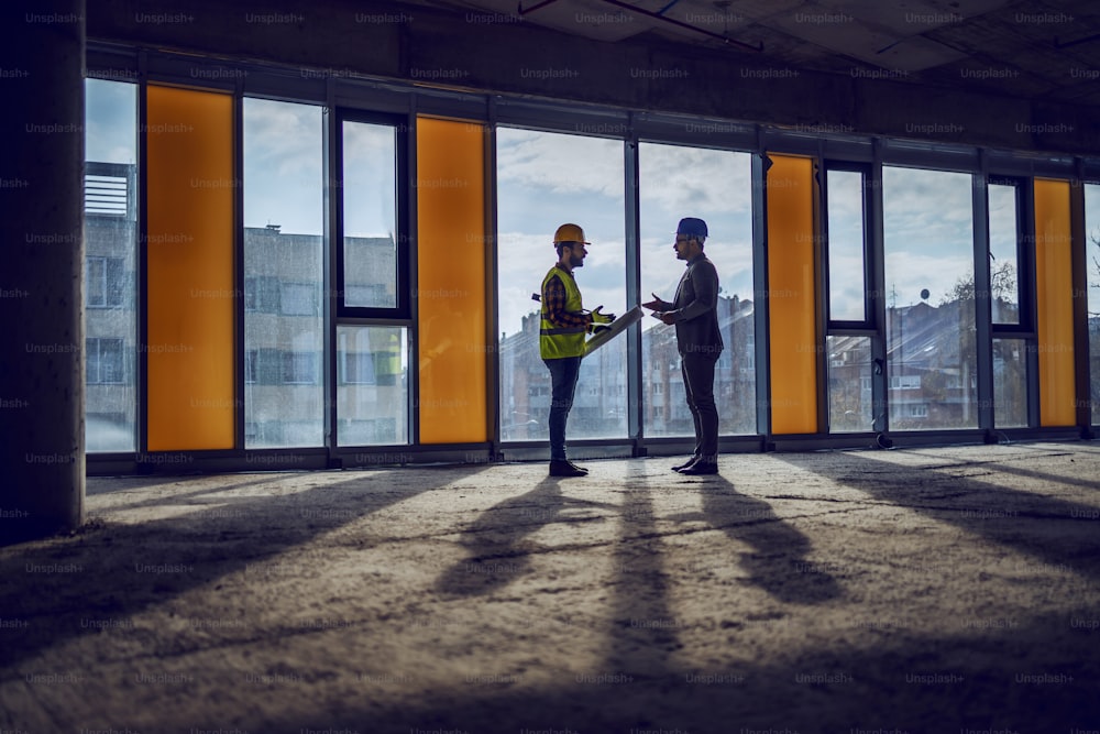 Silhouette of construction worker and architect standing near window in future business center and talking about realization of the project.