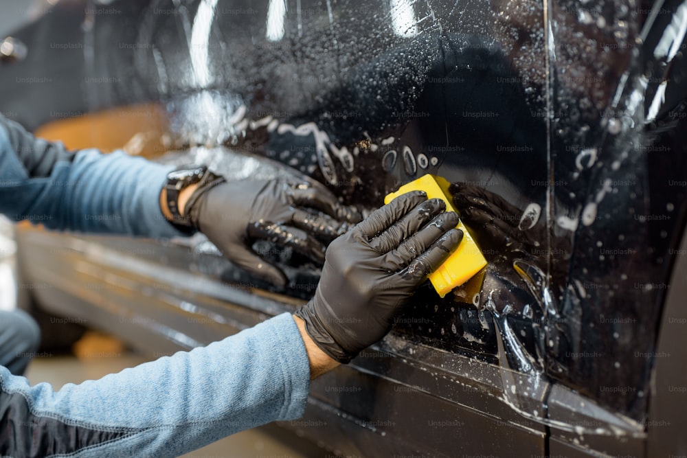 Worker sticking anti-gravel film on a car body with scrapper at the detailing vehicle workshop, close-up. Concept of car body protection with special films