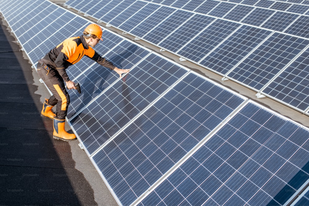 Well-equipped worker in protective orange clothing installing or replacing solar panels on a photovoltaic rooftop plant. Concept of maintenance and installation of solar stations