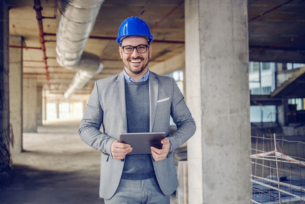Handsome cheerful caucasian unshaven architect in suit, with helmet on head and tablet in hands standing in building in construction process and looking at camera.