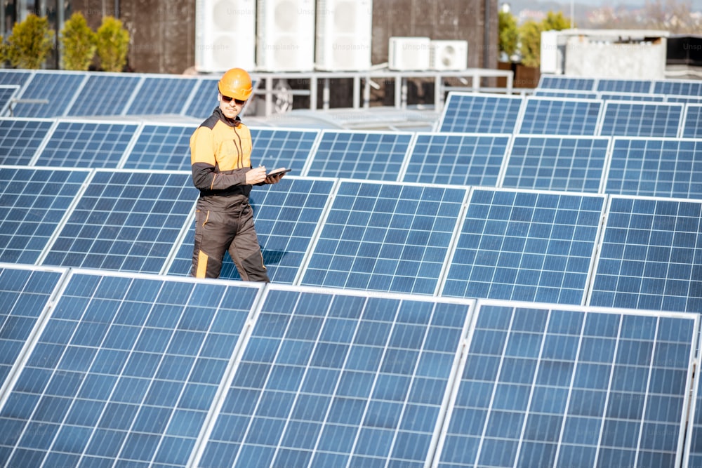 View on the rooftop solar power plant with engineer in protective workwear walking and examining photovoltaic panels. Concept of alternative energy and its maintenance