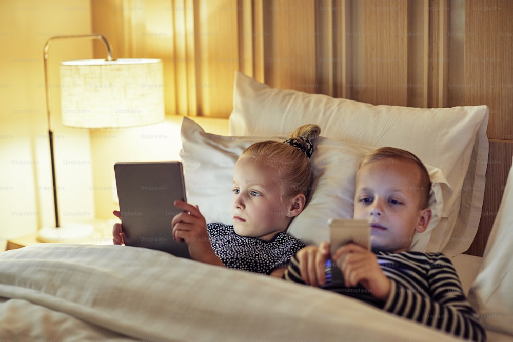 Cute little brother and sister lying in bed together using a digital tablet and smartphone before bedtime