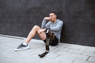 Joven deportista caucásico atractivo con pierna artificial sentado en el suelo, apoyado en la pared y comiendo manzana fresca.