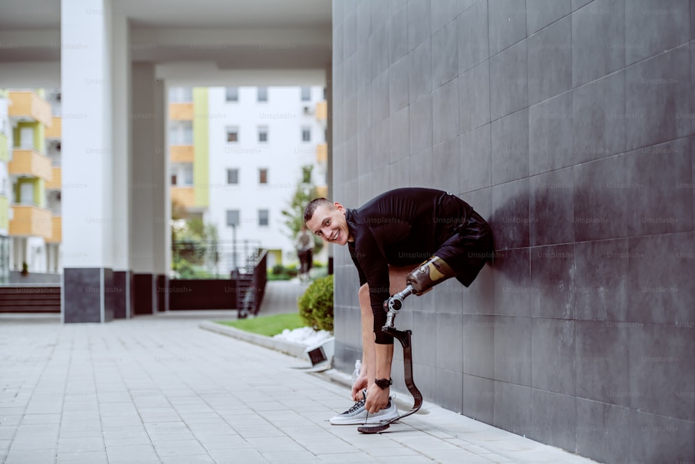 Vista lateral de un atractivo deportista caucásico con una pierna artificial apoyada en la pared y atándose el cordón de los zapatos.