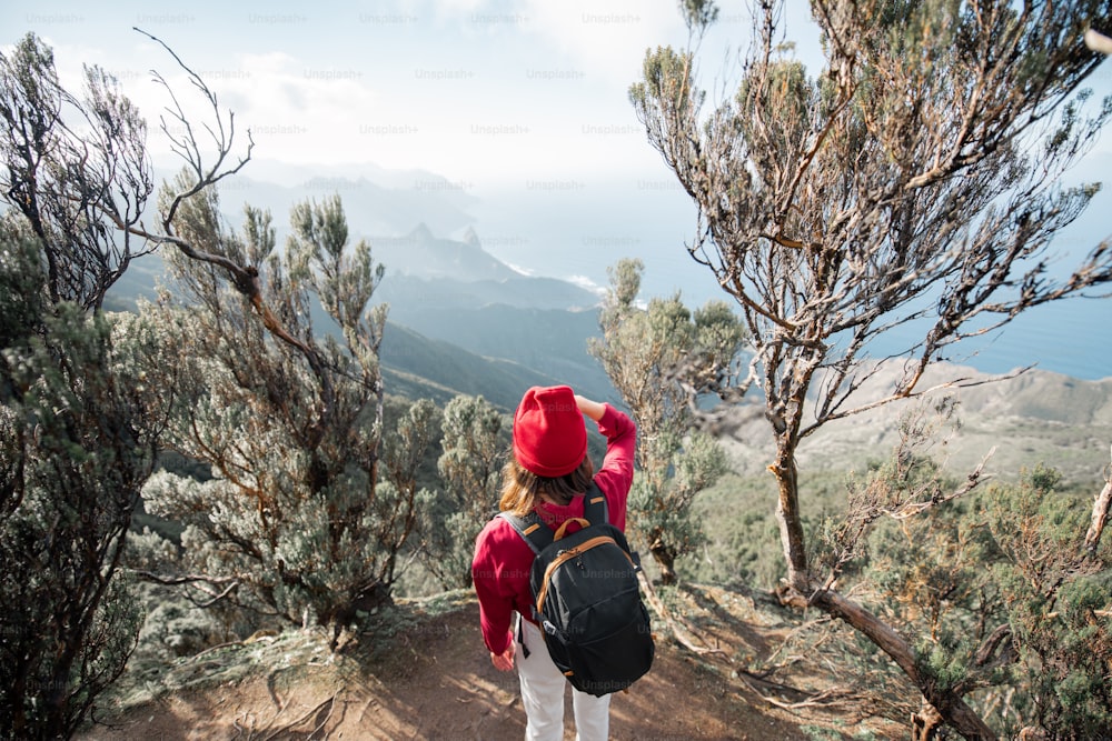 빨간 모자를 쓴 여자는 열대 우림의 산에서 높은 산을 여행하는 동안 테네리페 섬의 바위 해안선에서 숨막히는 전망을 즐기고 있다