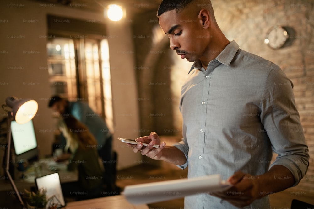 Imprenditore afroamericano che legge il messaggio sullo smartphone mentre lavora su rapporti aziendali di notte in ufficio.