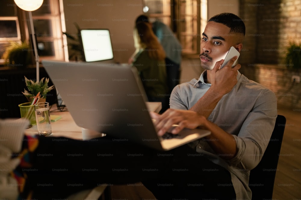 Empresario afroamericano que usa una computadora portátil y habla por teléfono móvil mientras trabaja hasta tarde en la oficina.
