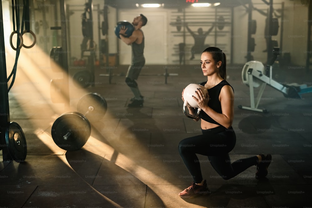Mulher atlética fazendo agachamentos divididos enquanto se exercita com kettle bell em uma academia.