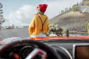 Portrait de style de vie d’une femme insouciante vêtue d’un pull et d’un chapeau colorés assis sur le capot de la voiture, profitant d’un voyage en voiture sur la route de montagne, vue à travers le pare-brise