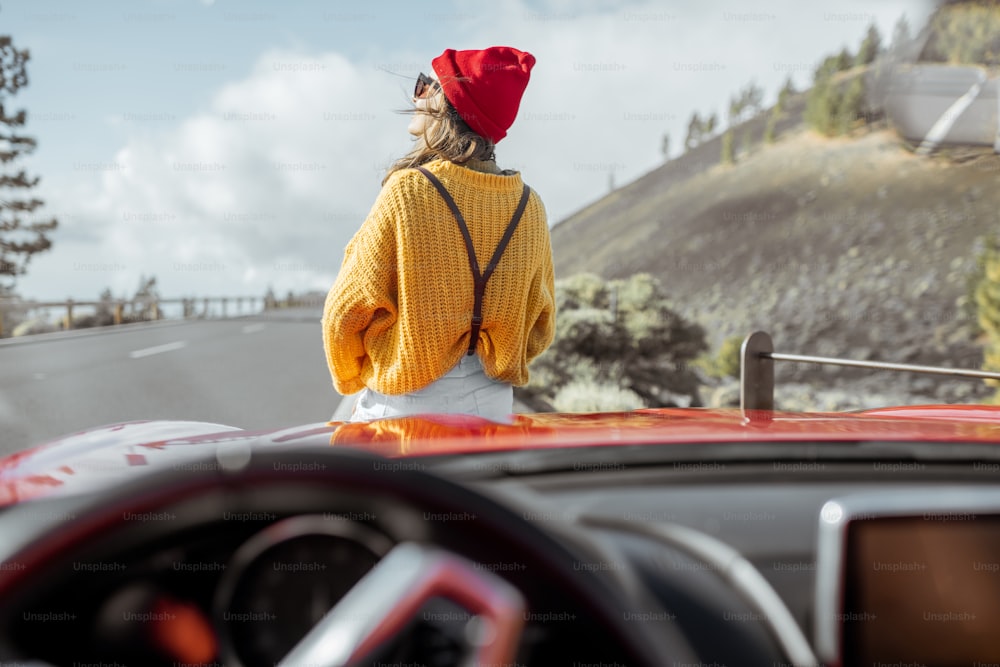 Lifestyle portrait of a carefree woman dressed casually in bright sweater and hat sitting on the car hood, enjoying road trip on the mountain road, view through the windshield