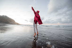 Lifestyle portrait of a carefree woman dressed in red shirt and hat walking on the beach at dusk. Wellness, happiness and life enjoyment concept