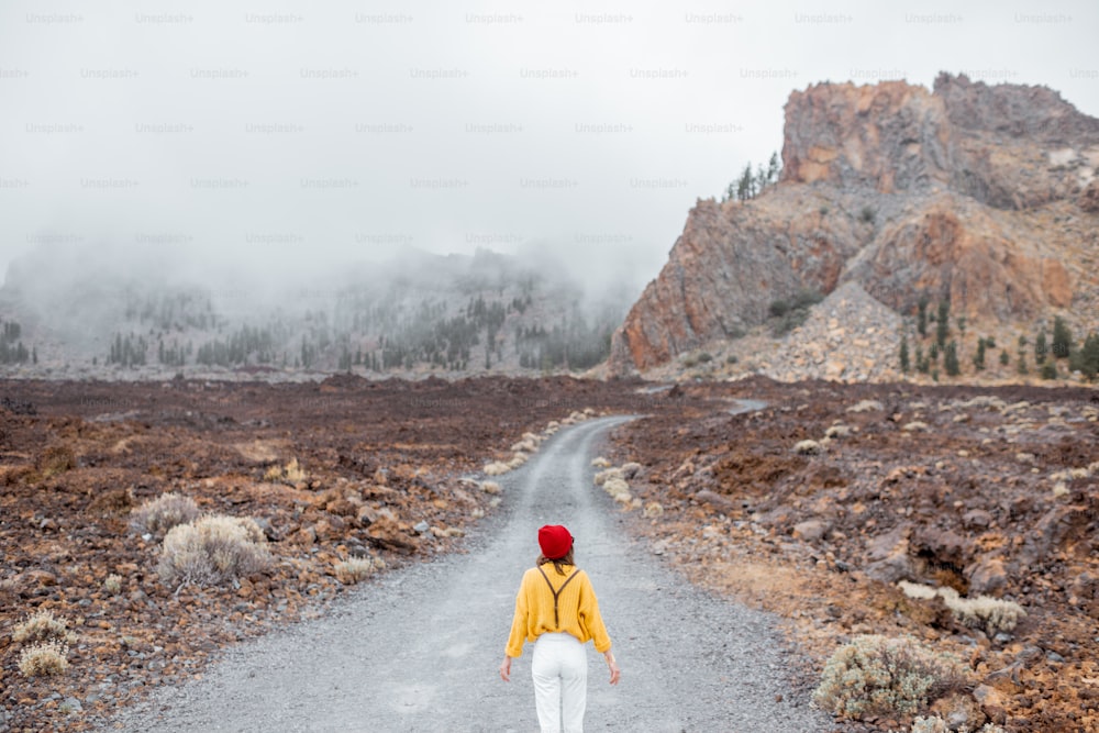 Picturesque rocky mountains in the clouds with dirt road on the volcanic valley. Woman in bright clothes walking on the road, wide view