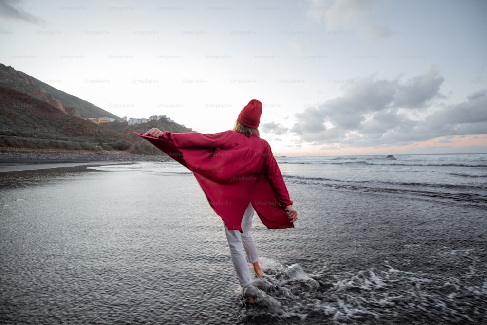 Lifestyle portrait of a carefree woman dressed in red shirt and hat walking on the beach at dusk. Wellness, happiness and life enjoyment concept