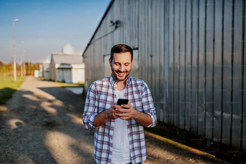 Young smiling unshaven caucasian farmer standing next to barn and using smart phone for texting.