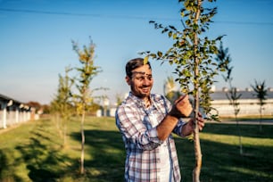 Young smiling happy agronomist standing in orchard and looking at fruit tree.