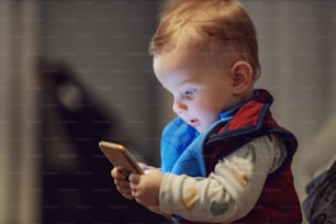 Adorable amazed little blond boy sitting in mother's lap and holding smart phone. He is surfing on internet.