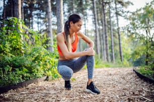 Young slim sportswoman crouching on footpath in nature and holding her injured knee.