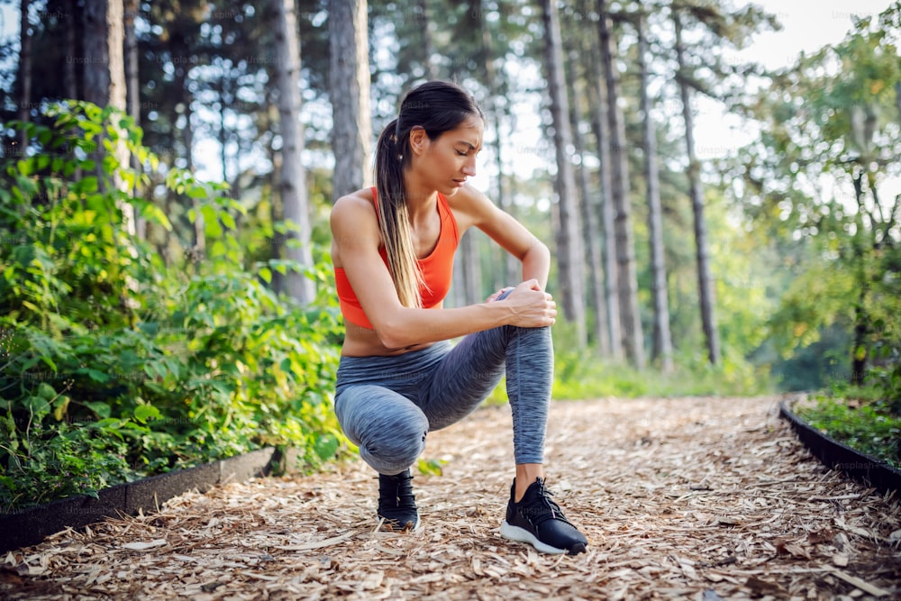 Young slim sportswoman crouching on footpath in nature and holding her injured knee.