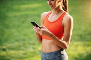 Young attractive slim sportswoman taking a break after running and using smart phone for texting messages on social media.
