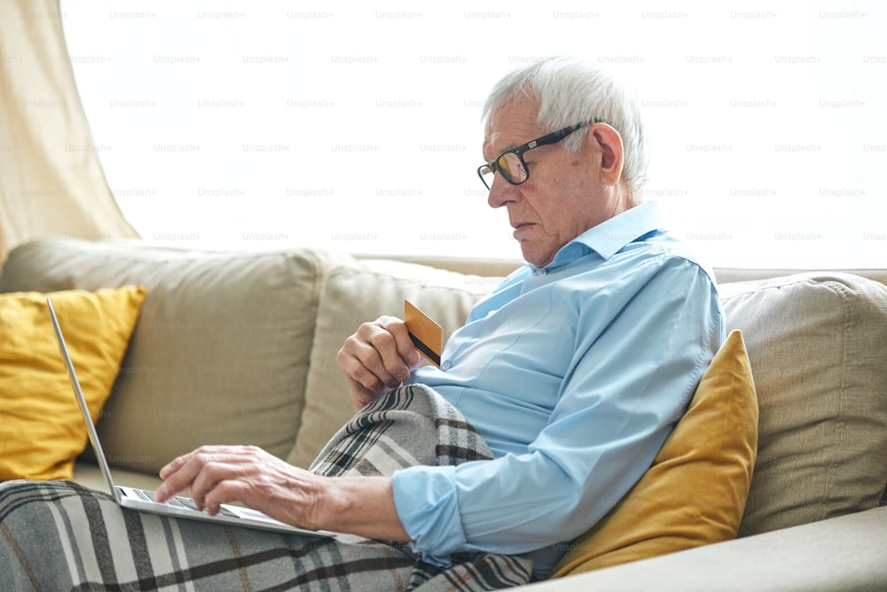 Retired grey-haired man wrapped in plaid relaxing on couch and ordering goods from online shop in front of laptop