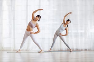 In voller Länge stehen zwei fitte, schlanke, attraktive junge Frauen mit gespreizten Beinen und Händen in den Hüften auf der Matte im Yogastudio.