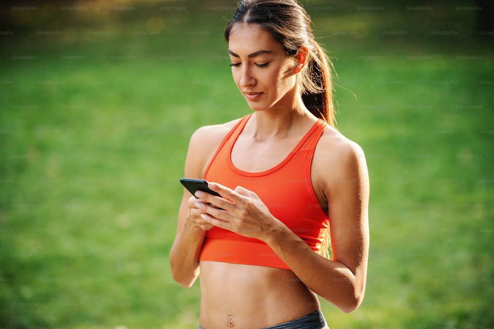 Portrait of young attractive slim sportswoman taking a break after running and using smart phone for texting messages on social media.